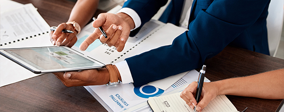 Two consultants reviewing marketing statistics on a tablet and printed reports at a desk.